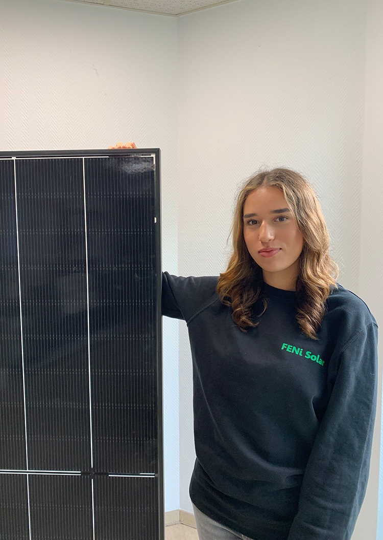 Eine Frau mit lockigem Haar und schwarzem FENESolar-Pullover steht neben einem Solarmodul, mit ruhigem Gesichtsausdruck.